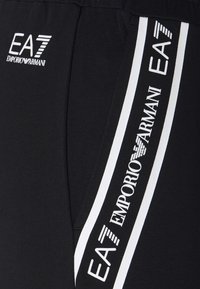 Černé atletické šortky s bílými bočními pruhy a textem "EA7 EMPORIO ARMANI". Vyrobené z měkké tkaniny, vhodné na sport a volnočasové nošení.