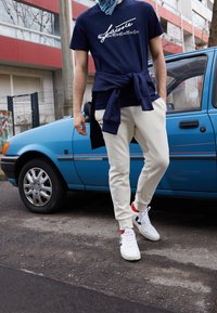 Blue car parked on the street; person wearing a navy T-shirt with white print, beige trousers, and white sneakers with red accents.