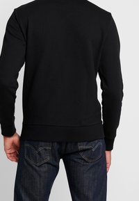 Felpa nera con maniche lunghe, polsini e orlo a coste. Abbinata a jeans in denim scuri con cuciture sottili e dettagli sulla tasca posteriore.