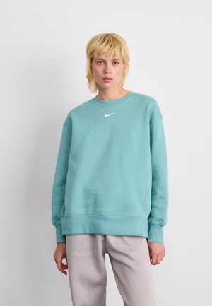 En blond person som har på seg en teal Nike hettegenser og lysegrå bukser, står mot en enkel hvit bakgrunn.
