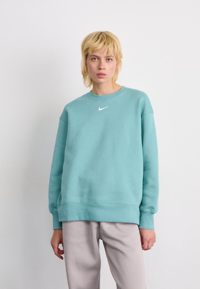 Persona rubia vestida con una sudadera de Nike color aguamarina y pantalones gris claro, de pie frente a un fondo blanco liso.