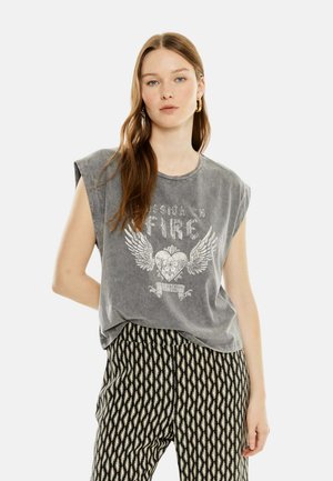 Joven con cabello largo lleva una camiseta gris sin mangas con gráfico y pantalones estampados en blanco y negro, de pie con una mano detrás de la espalda.