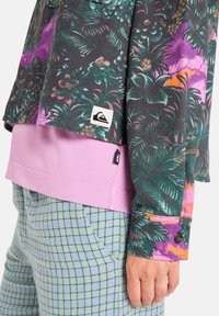 Gemusterte Jacke mit grünem, lila und pinkem Blattdesign; über einem pinken Shirt getragen; kombiniert mit hellblauen karierten Hosen. Enthält Logo-Tags.