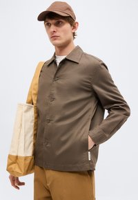 Άνδρας που φορά καφέ μπουφάν, ταιριαστό καπέλο, καφέ παντελόνια και κρατά μια μπεζ και καφέ τσάντα ώμου, κοιτάζει πλάγια.