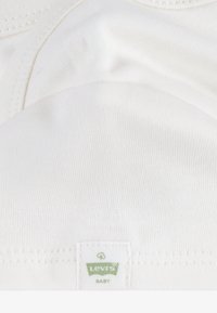 Bijeli pamučni materijal s glatkom teksturom; sadrži etiketu s natpisom "Levi's" i "BABY" u zelenoj boji. Jednostavan, minimalistički dizajn.