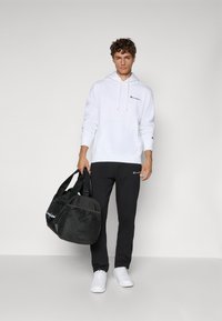 White hoodie with drawstrings and logo, black sweatpants with logo, white sneakers, and a black duffel bag with handles, textured fabric.