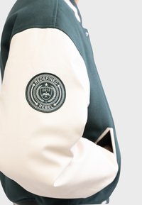 Green and cream varsity-style jacket with a textured fabric body, smooth leather sleeves, and a round embroidered patch on the arm.
