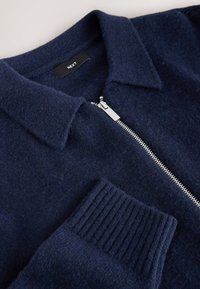 Granatowy sweter na zamek wykonany z miękkiego materiału, z kołnierzykiem, prążkowanymi mankietami oraz metalowym zamkiem błyskawicznym z przodu.