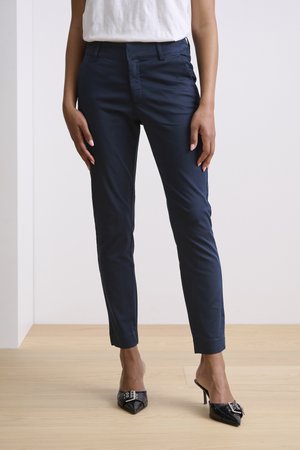 Tamsiai mėlynos slim-fit kelnės su lygia tekstūra, su šoniniais kišenėmis ir plokščiu priekiu, derinamos su juodais smailiais aukštakulniais.
