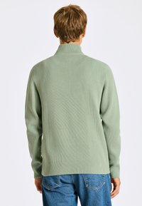 Ljusgrön ribbad stickad tröja med hög krage, långa ärmar och sidsprund, i kombination med blå denimjeans.