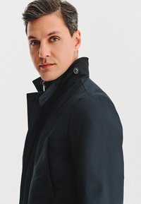 Czarny płaszcz z wysokim kołnierzem, o smukłych liniach i guzikowych akcentach. Gładka tekstura na modelu na jednolitym tle.