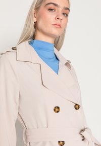 Heller beiger Trenchcoat mit doppelt geknöpftem Verschluss, großen Schildpattknöpfen und einem taillierten Gürtel. Schmale Revers mit Schulterklappen.