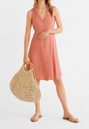Femme portant une robe corail sans manches à boutons sur le devant, tenant un grand sac à main beige tressé, portant des sandales beiges et des bijoux en or.