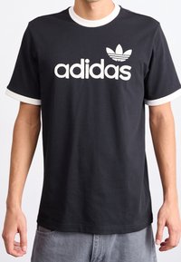 Musta puuvillainen t-paita, jossa on valkoinen ribbikangas kauluksessa ja hihojen päissä. Etupuolella on suuri valkoinen "adidas"-logo ja -symboli vasemmassa rinnassa.
