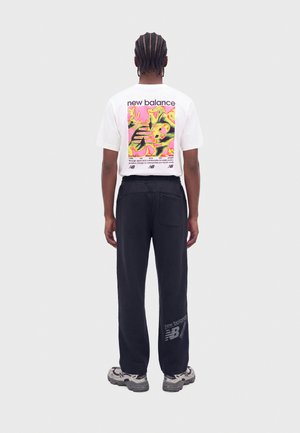 Pessoa com cabelo trançado a usar uma t-shirt branca com gráfico da New Balance, calças pretas com logótipo e ténis cinzentos, em pé de costas.