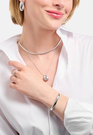 Silberschmuck bestehend aus einem Choker, einer mehrlagigen Halskette mit einem tropfenförmigen Anhänger, Creolen und einem glatten Armreif. Getragen zu einer weißen Bluse.