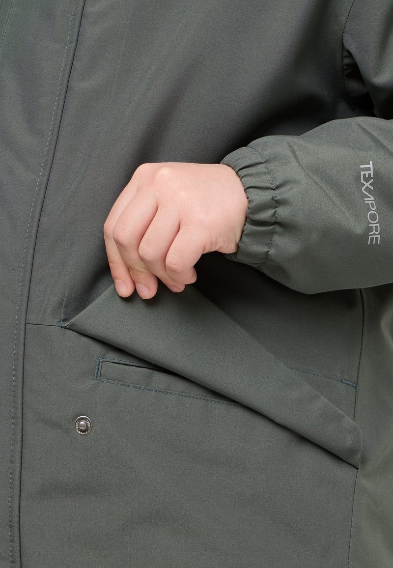 Dunkelgrüne TXAPORE-Jacke mit elastischen Bündchen, in der eine Hand in eine Seiten tasche greift, die eine elegante, glatte Textur zeigt.