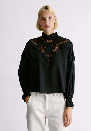 Blusa nera con colletto arricciato, motivo in pizzo sul petto e maniche lunghe. Abbinata a pantaloni bianchi, con una texture di tessuto liscia.