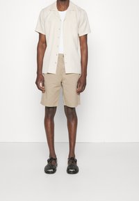 Košile s krátkým rukávem v béžové barvě s jednou náprsní kapsou, zkombinovaná s khaki šortkami. Černé sandály na pásky dotvářejí celkový outfit.