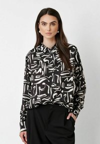 Woman with long dark hair wears black and white abstract patterned button-up shirt and black pants, standing with one hand in pocket.