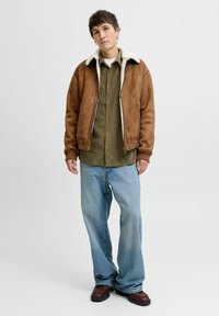Una giovane persona è in piedi indossando una giacca di shearling marrone, una camicia a bottoni verde oliva, jeans leggeri azzurri larghi e scarpe in pelle marrone su uno sfondo bianco.