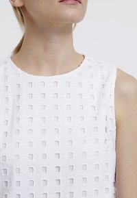 Haut blanc sans manches avec un design texturé comprenant des découpes carrées, un tissu lisse et un col rond.