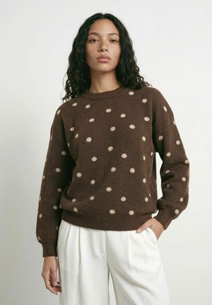 Femme aux longs cheveux bouclés portant un pull marron à pois blancs et un pantalon blanc, debout devant un fond clair uni.