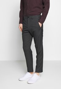 Pantaloni grigi su misura con una texture liscia, dotati di tasche laterali e una vestibilità elegante, abbinati a sneakers bianche e una camicia a quadretti.