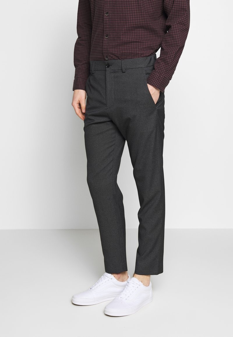 Pantaloni grigi su misura con una texture liscia, dotati di tasche laterali e una vestibilità elegante, abbinati a sneakers bianche e una camicia a quadretti.