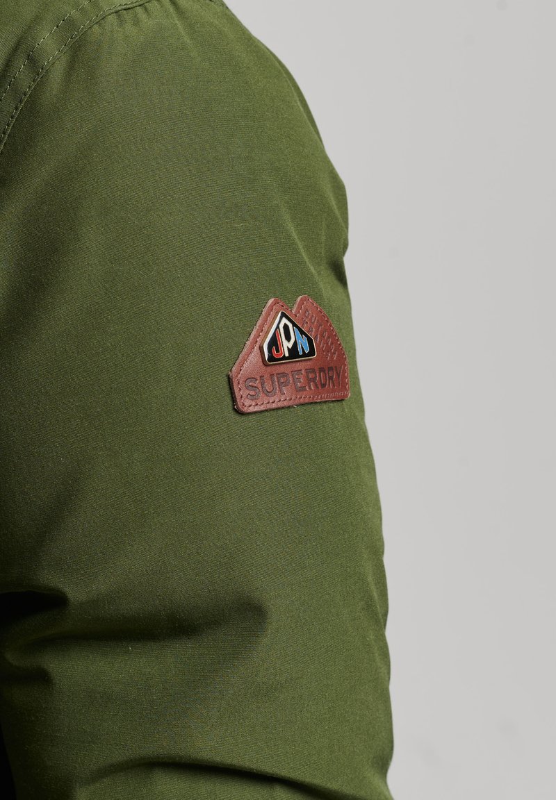 Olivenfarbene Jacke mit glatter Textur und einem braunen Lederpatch am Arm, das ein dreieckiges Design und ein Logo zeigt.