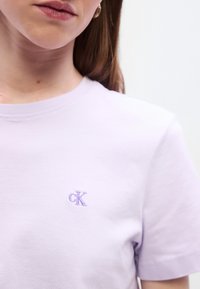 Ελαφριά βαμβακερή μπλούζα σε ανοιχτό λιλά χρώμα με στρογγυλή λαιμόκοψη και κοντά μανίκια, διακοσμημένη με μικρό κεντημένο λογότυπο "ck" σε μωβ χρώμα στην αριστερή πλευ ρά του στήθους.