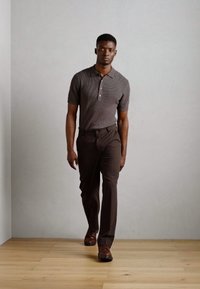 Polo gris rayé à manches courtes avec trois boutons, associé à un pantalon marron ajusté et des chaussures marron, se tenant sur un sol en bois.