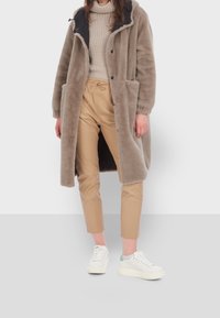 Dlouhý, světle šedý kožešinový kabát s tmavým límcem, spárovaný s béžovými zúženými kalhotami a bílými teniskami se světle modrým akcentem. Detaily texturované látky.