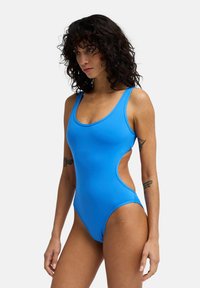 Ολόσωμο μπλε μαγιό με φαρδιές τιράντες, κοψίματα στα πλάγια και λεία υφή. Διαθέτει χαμηλή πλάτη και ψηλές τιράντες στα πόδια.