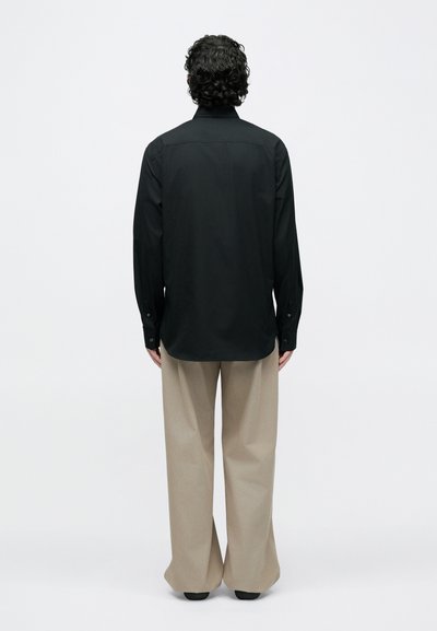 Mand med krøllet hår iført en sort langærmet skjorte og beige bukser med vide ben, stående med ryggen mod en hvid baggrund.