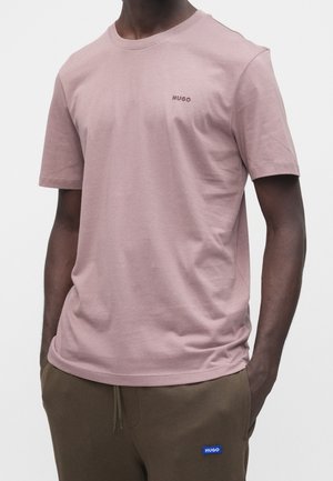 Mand iført en lys lilla Hugo t-shirt og brune sweatpants med et blåt Hugo-mærke, stående foran en hvid baggrund.