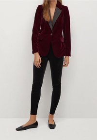 Bordeauxrode fluwelen blazer met een zwarte satijnen revers, twee voorzakken, gecombineerd met glanzende zwarte leggings en platte zwarte schoenen.