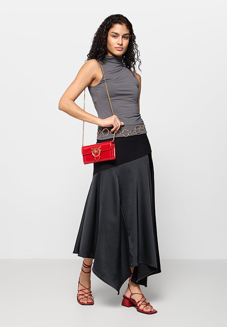 Szary bezrękawnik, czarna asymetryczna spódnica, czerwona lakierowana torebka z złotymi akcentami oraz czerwone sandały na cienkich paskach. Gładkie tekstury, elegancki design.