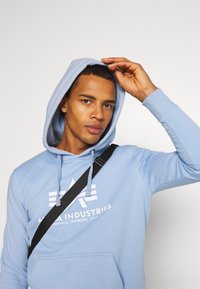 Felpa leggera azzurra con coulisse e tasca frontale. Logo stampato in bianco. Il modello indossa una borsa a tracolla nera.