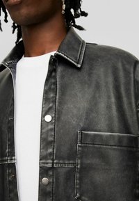 Černá bunda-košile z uměl�é kůže s špičatým límečkem, stříbrnými zatrhávacími knoflíky a náprsní kapsou. Má hladkou texturu a strukturovaný střih.