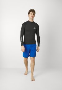 Langermet svart rash guard med logo på brystet, kombinert med blå svømmetights. Figuren står barføtt på en nøytral bakgrunn.