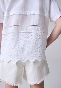 Witte blouse met geborduurde kanten zoom en uitsnijdingen, gecombineerd met ecru denim shorts met rafelige randen. Subtiele textuur en elegant ontwerp.