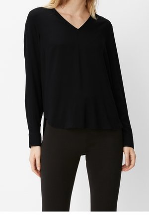 Kvinde iført en sort langærmet V-hals bluse kombineret med sort tætsiddende bukser, stående mod en ensartet lys baggrund.