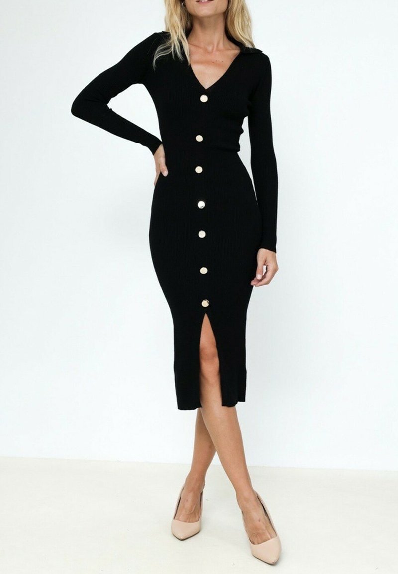 Robe noire côtelée avec un décolleté en V, des manches longues et des boutons sur le devant. Présente une fente jusqu'au genou pour plus de liberté de mouvement et une silhouette près du corps.