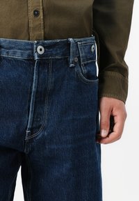 Jean en denim bleu foncé avec une coupe droite, comportant une taille standard, deux poches avant et des détails de couture contrastés.