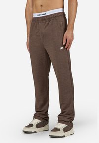 Brune teksturerte joggebukser med elastisk midje, prydet med et hvitt logo-patch og en avslappet passform, kombinert med kremfargede og brune sneakers.