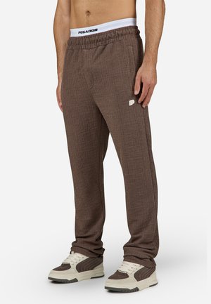 Brown textured jogger pants with an elastic waistband, featuring a white logo patch and a relaxed fit, paired with cream and brown sneakers.