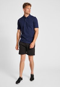 Marinblå pikétröja med korta ärmar, tvåknappsslutning och ribbad krage, kombinerad med svarta shorts och svarta sportskor.