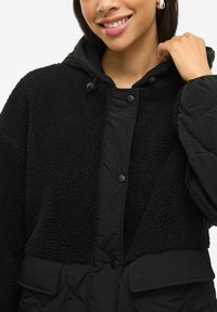 Melna jaka ar mīkstu flīsa ārpusi, quilted tekstūru, kapuci un pogu aizdari. Ietver priekšējos kabatas.