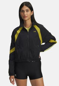 Zwarte windbreakerjas met gele diagonale strepen, ritssluiting aan de voorkant, twee zijzakken en een elastische tailleband. Lichtgewicht, soepel stof.